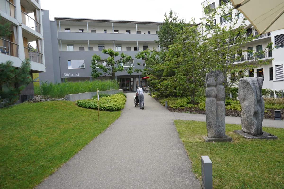 Park in der Zollinger Stiftung. Mann begleitet Frau in Rollstuhl.